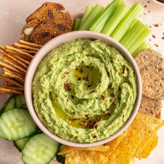 Overhead view of arugula hummus in a bowl with crackers and vegetables surrounding it.