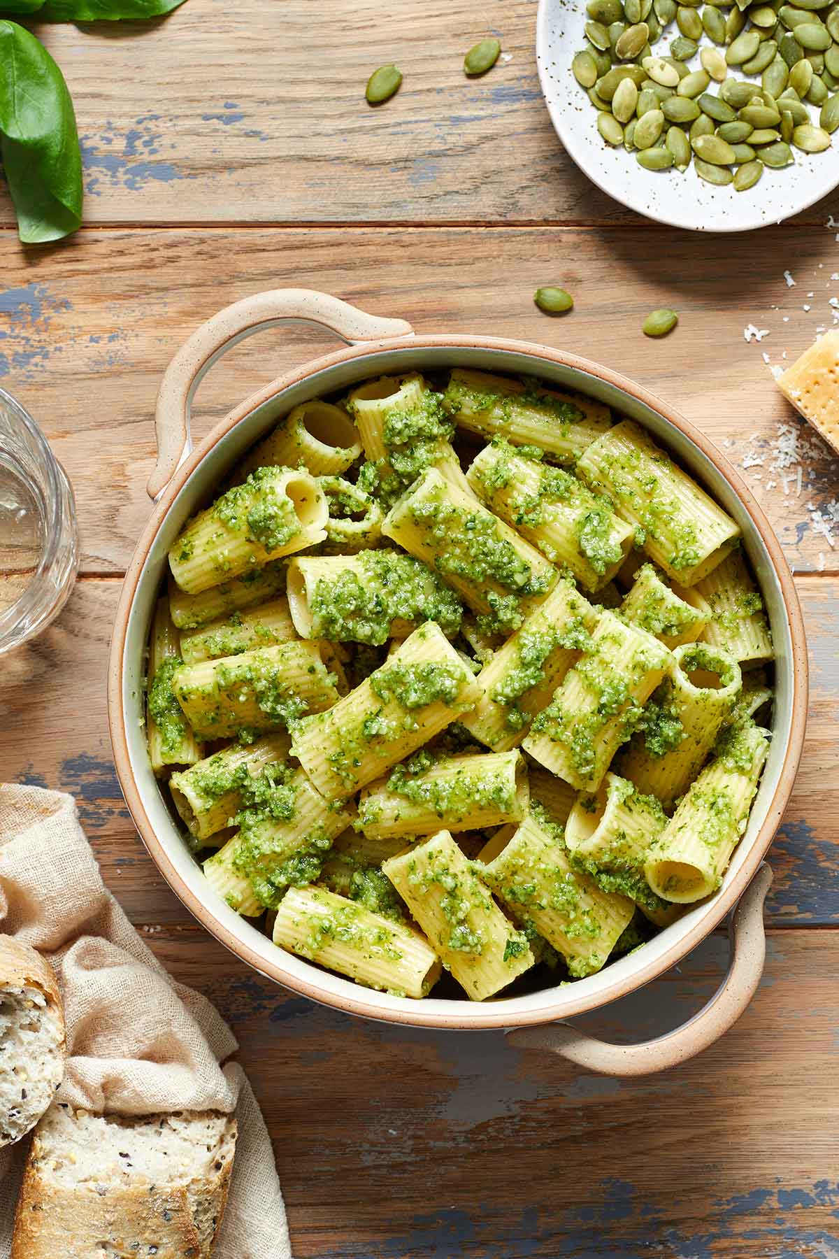 Pesto pasta in a bowl with crusty bread, cheese and pumpkin seeds on the side.