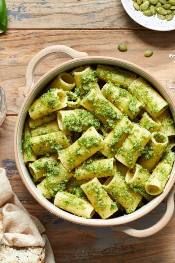 Pasta with pumpkin seed pesto in a bowl with a glass of wine, bread, pumpkin seeds and parmesan cheese surrounding it.
