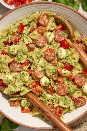 Pesto pasta salad with tomatoes, red peppers and mozzarella in a white bowl with wooden salad servers.