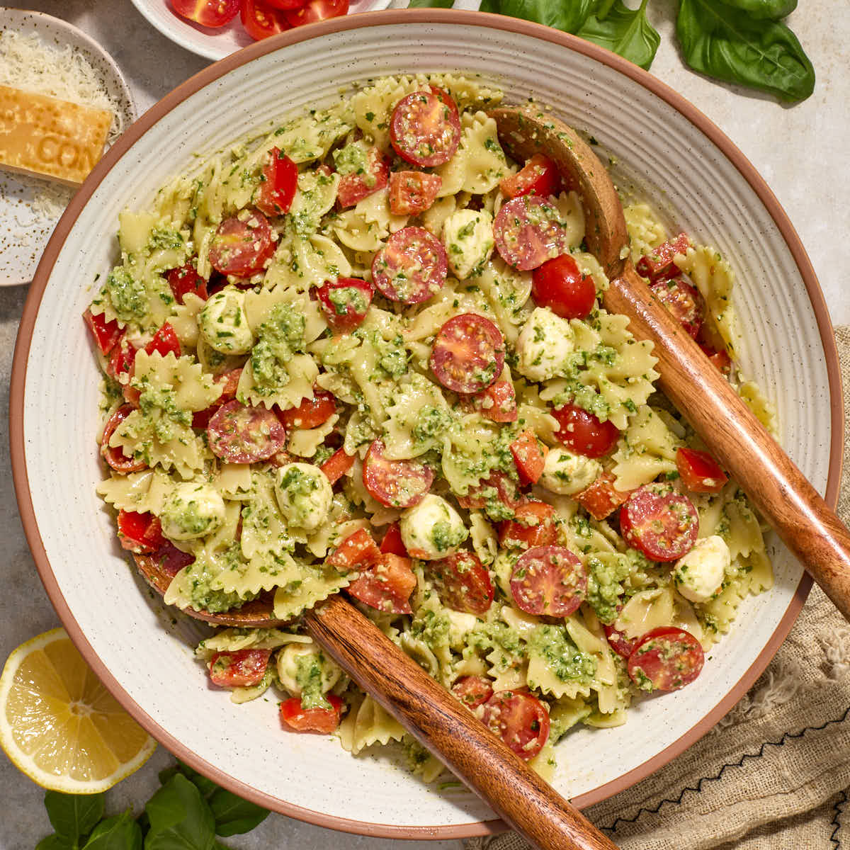 Pesto pasta salad with tomatoes, red peppers and mozzarella in a white bowl with wooden salad servers.