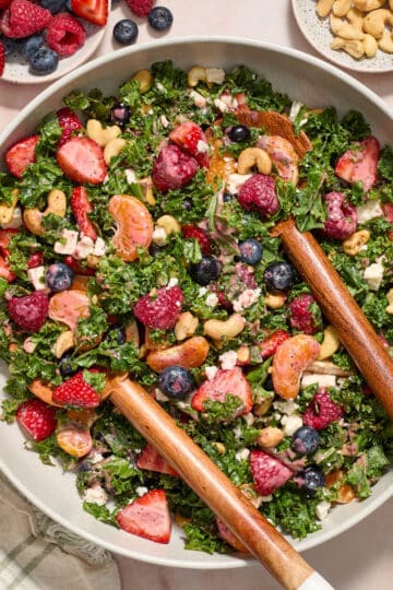 Kale berry citrus salad in a large serving bowl with wooden serving spoons inserted into it.