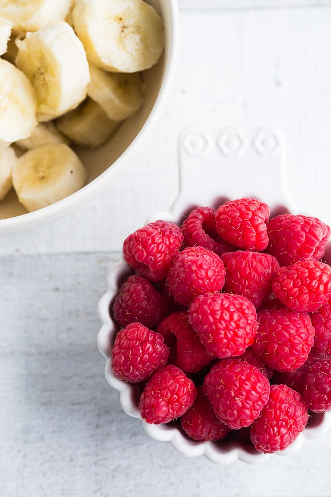 Overhead view of bowls of raspberries and chopped bananas on a white wooden surface.