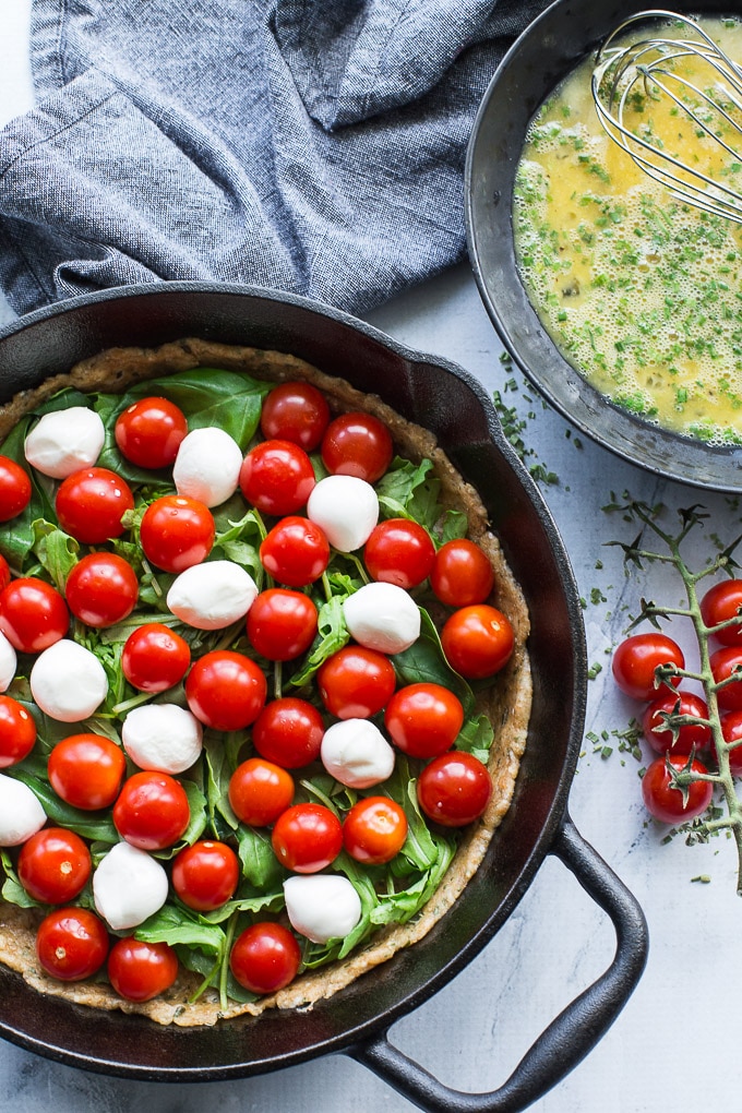 Overhead view of Tomato, Arugula and Mozzarella Quiche being assembled in a cast iron pan. A bowl of scrambled eggs next to it.