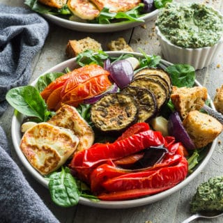 Up-close view of two plates of Salad with Roasted Vegetables and Halloumi Cheese next to a cup of pesto on a wooden surface.