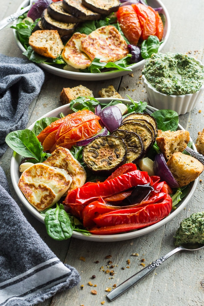 Up-close view of two plates of Salad with Roasted Vegetables and Halloumi Cheese next to a cup of pesto on a wooden surface.