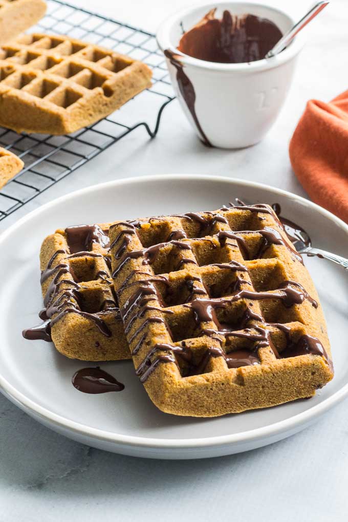 Side view of Grain-Free Pumpkin Chocolate Chip Waffles on a plate with waffles on a cooling rack and a dish of melted chocolate in the background.