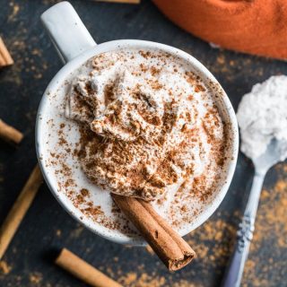 Overhead view of a Pumpkin Spice Collagen Latte on a dark surface surrounded by cinnamon sticks.