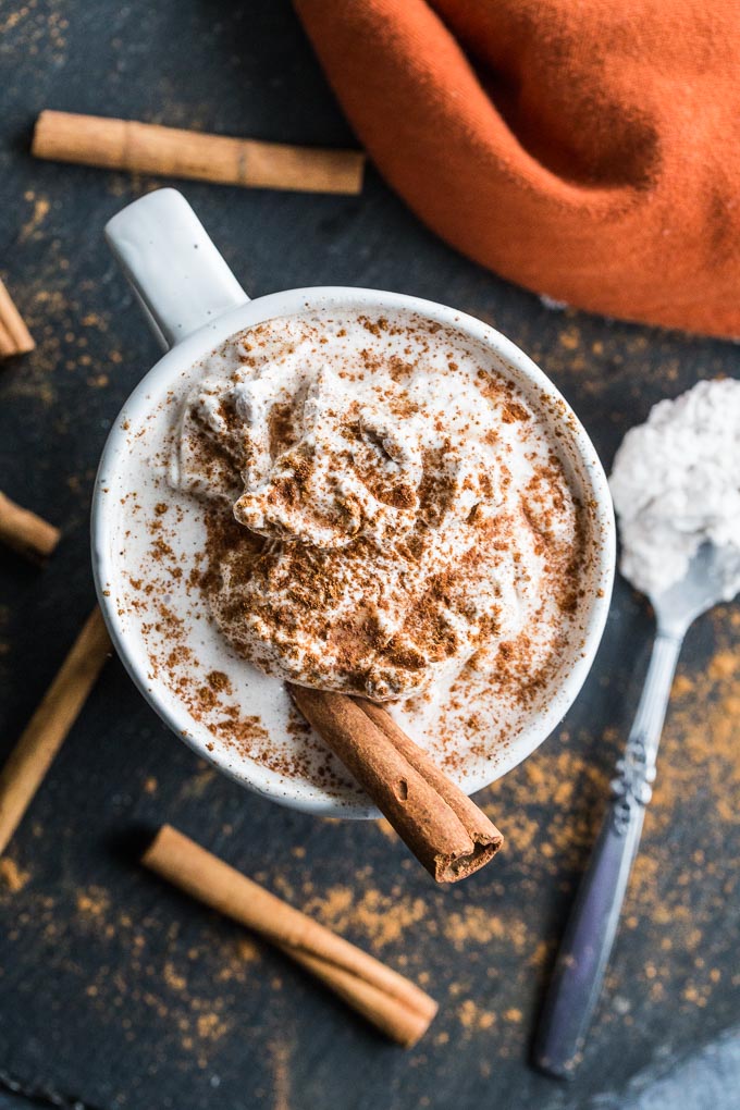 Overhead view of a Pumpkin Spice Collagen Latte on a dark surface surrounded by cinnamons sticks.