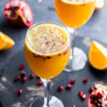 Two Ginger Beer Mimosas in wine glasses on a dark surface, surrounded by pomegranate arils.