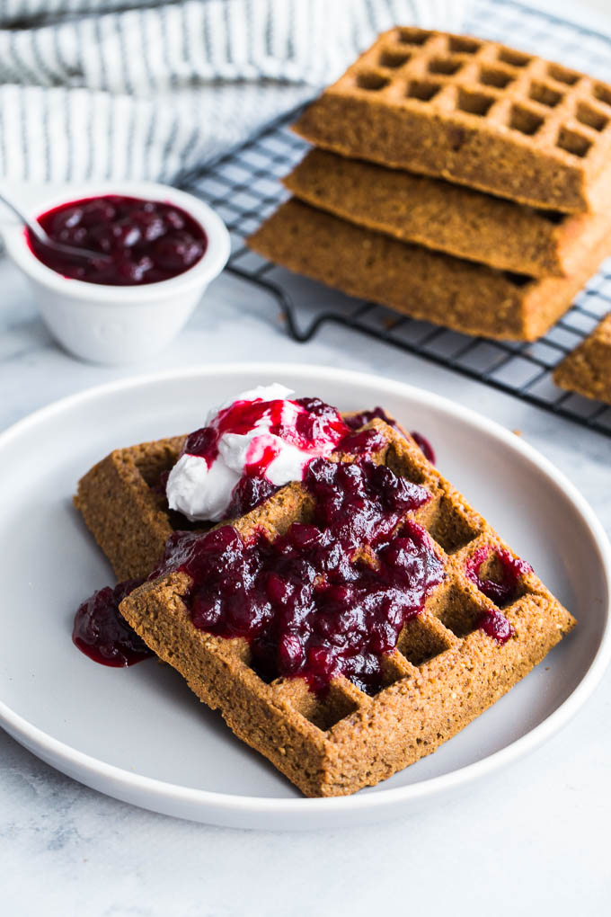 Two Gingerbread Waffles with Partridgeberry Compote on a grey plate, with a stack of waffles in the background.