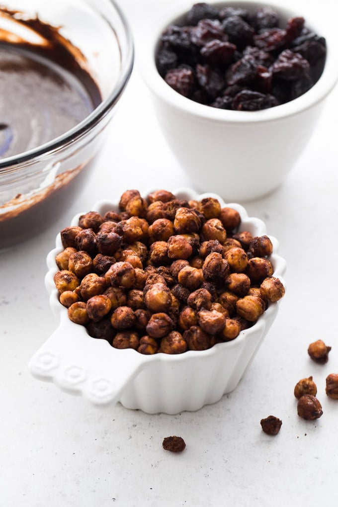 Up-close view of roasted chickpeas in a white cup with raisins and melted chocolate in the background.