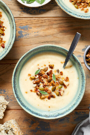 Parsnip and pear soup served in green bowls on a wooden table with savoury granola on top of the soup.