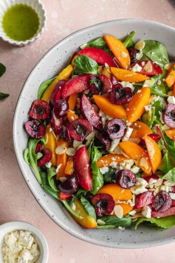 Overhead of stone fruit salad in a grey bowl with vinaigrette and other toppings on the side.