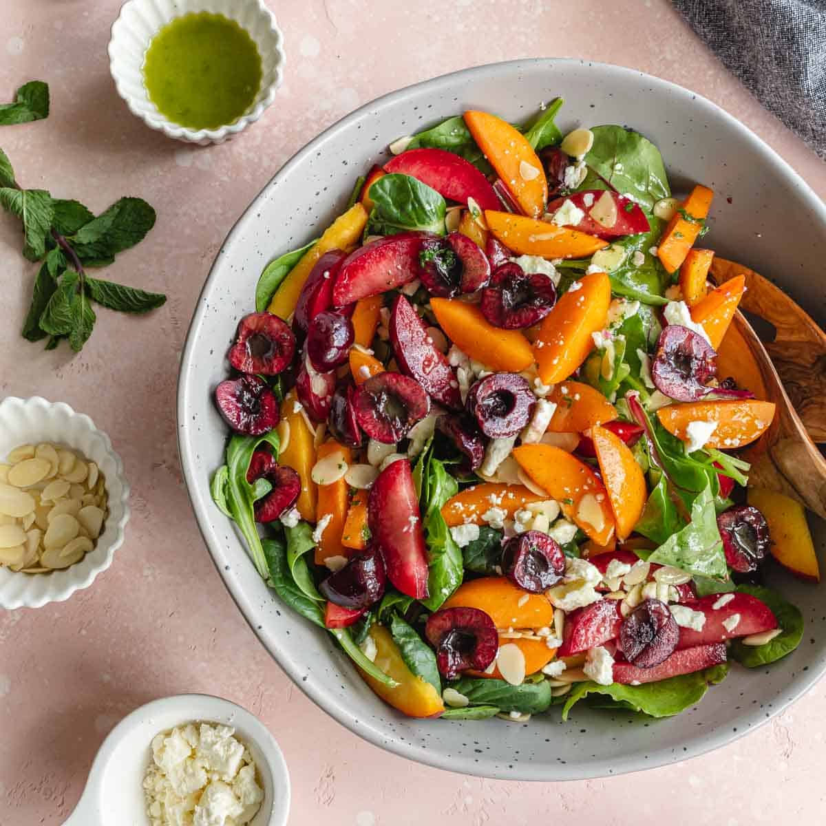 Overhead of stone fruit salad in a grey bowl with vinaigrette and other toppings on the side.