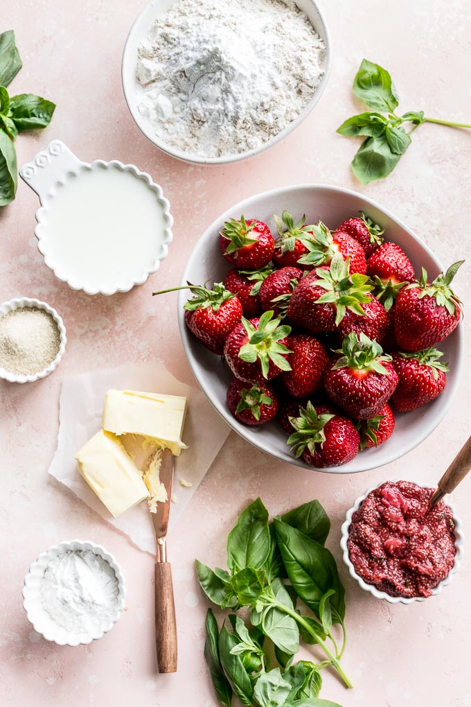 Overhead view of ingredients to make strawberry basil cobbler arranged on a pink surface.