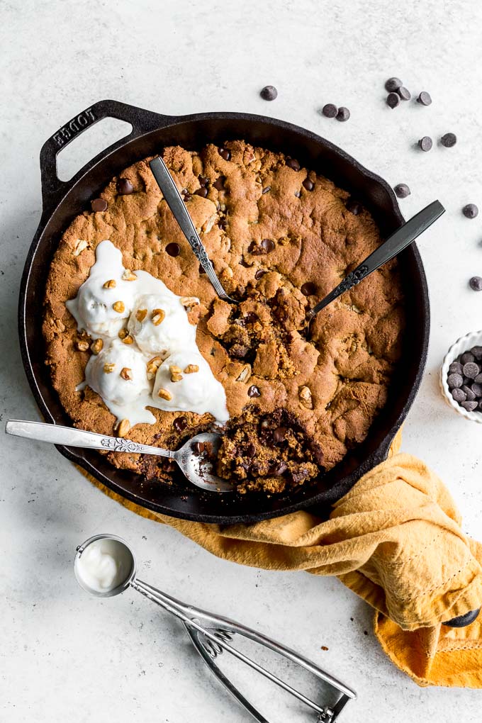 Pecan chocolate chip skillet cookie with melting ice cream on top, three spoons inserted into it and chocolate chips off to the side.