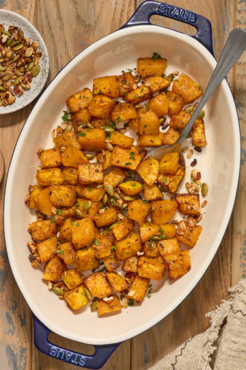 Air fryer butternut squash cubes in a serving dish with chopped pecans, pumpkin seeds and fresh herbs.