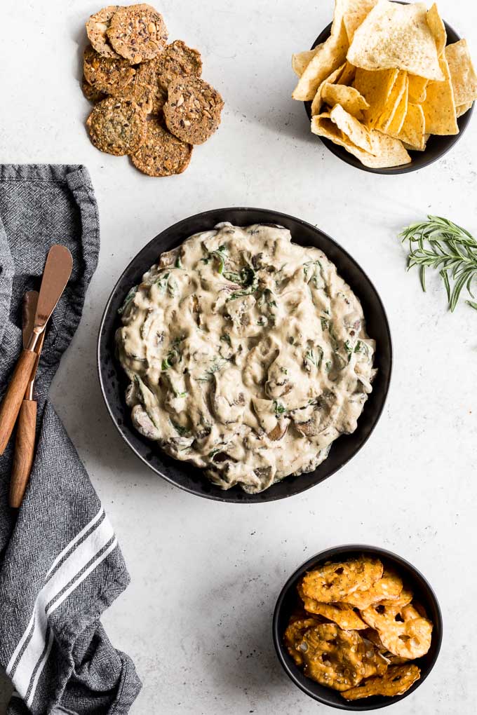 Overhead view of Mushroom Spinach Dip in a black bowl surrounded by small bowls of chips and pretzels.