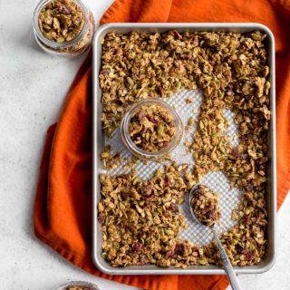 Pumpkin Spice Granola on a baking sheet with granola being spooned into glass jars.