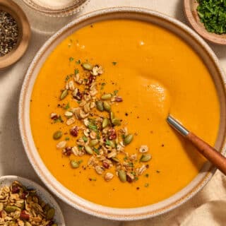 A bowl of roasted red pepper and butternut squash soup with toppings on the side.