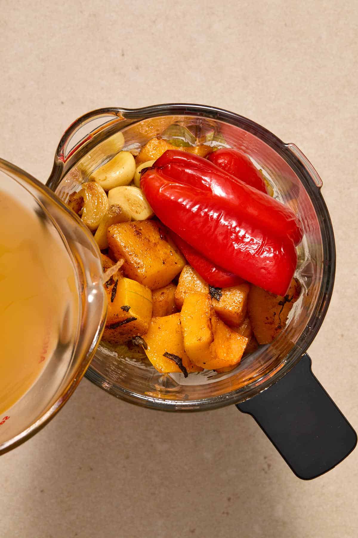 Roasted soup ingredients in a blender with vegetable broth being poured in.