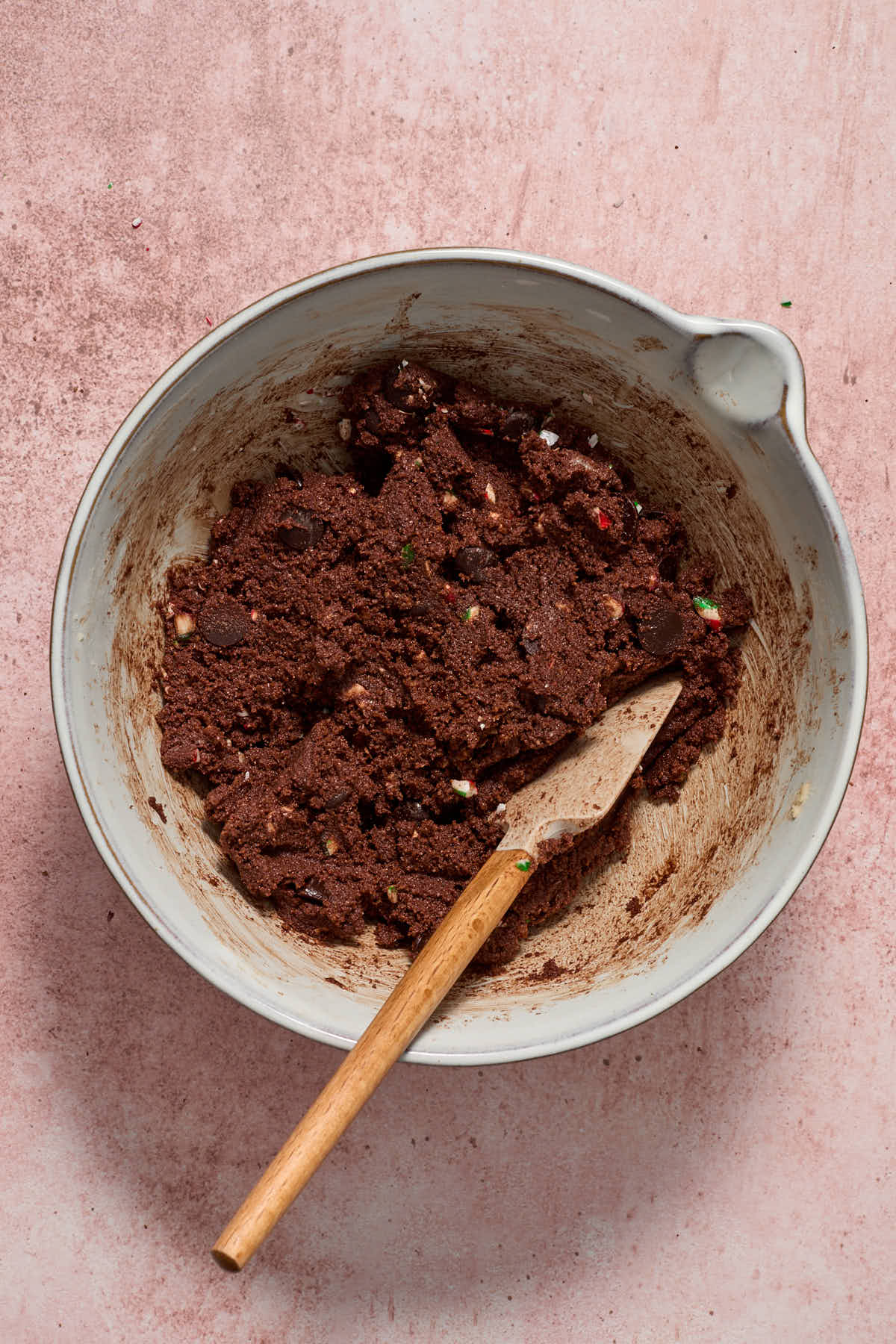 Cookie dough batter in a white bowl with candy cane pieces and chocolate chips mixed in with a spatula.