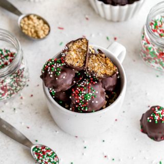 Crispy Peanut Butter Balls in a white mug on a white surface next to jars of sprinkles.