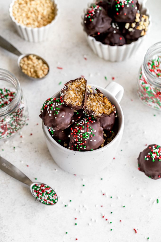 Crispy Peanut Butter Balls in a white mug on a white surface next to jars of sprinkles.