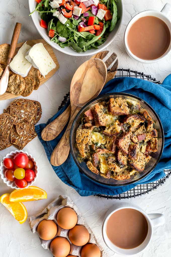 Overhead view of an egg strata in a glass dish surrounded by fruit, veggies, cheese and crackers.