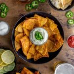 Overhead view of air fryer tortilla chips arranged in bowls with dips, beer and jalapenos surrounding it.
