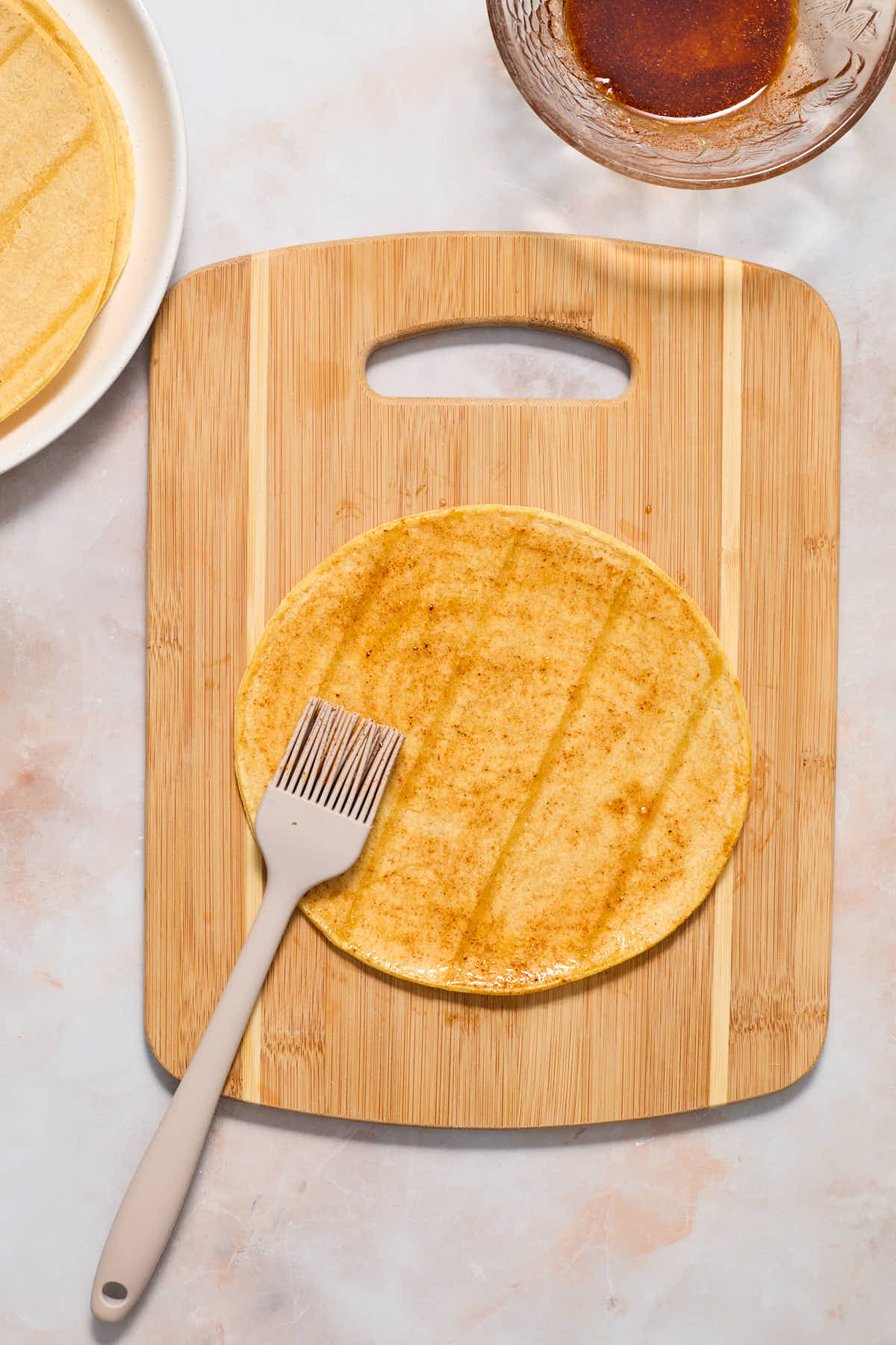 A corn tortilla on a wooden board being brushed with the oil mixture.