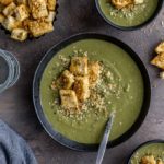 Bowls of green pea soup and croutons arranged on a grey surface.