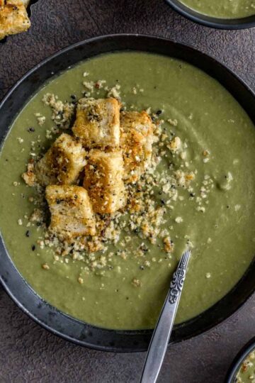 Overhead of green pea soup in a black bowl with croutons on top.