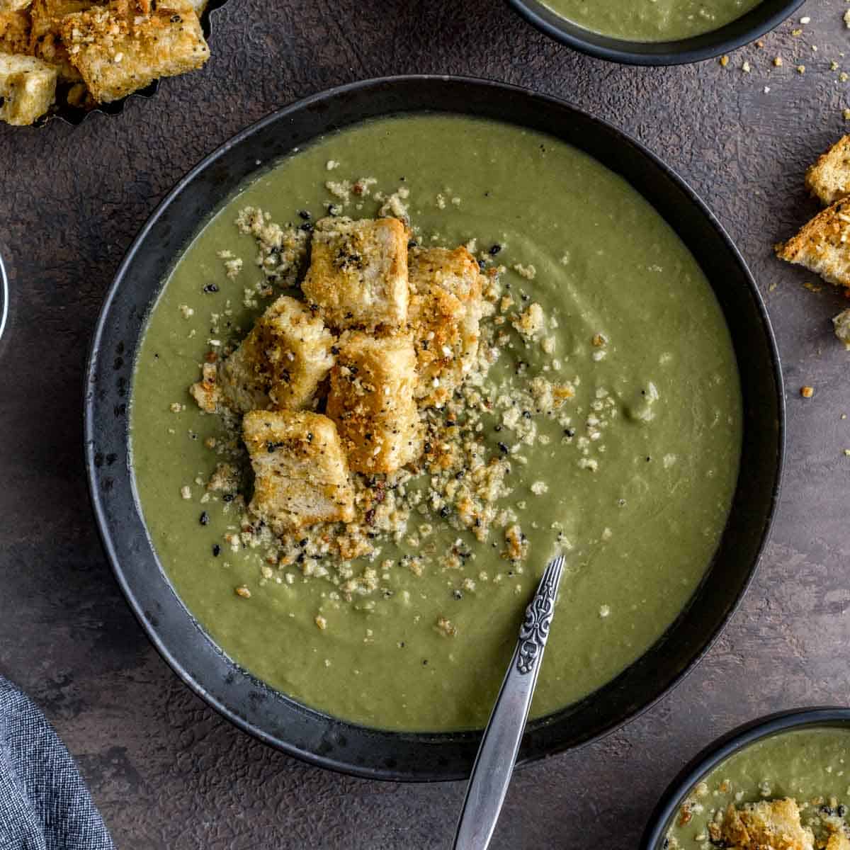 Overhead of green pea soup in a black bowl with croutons on top.