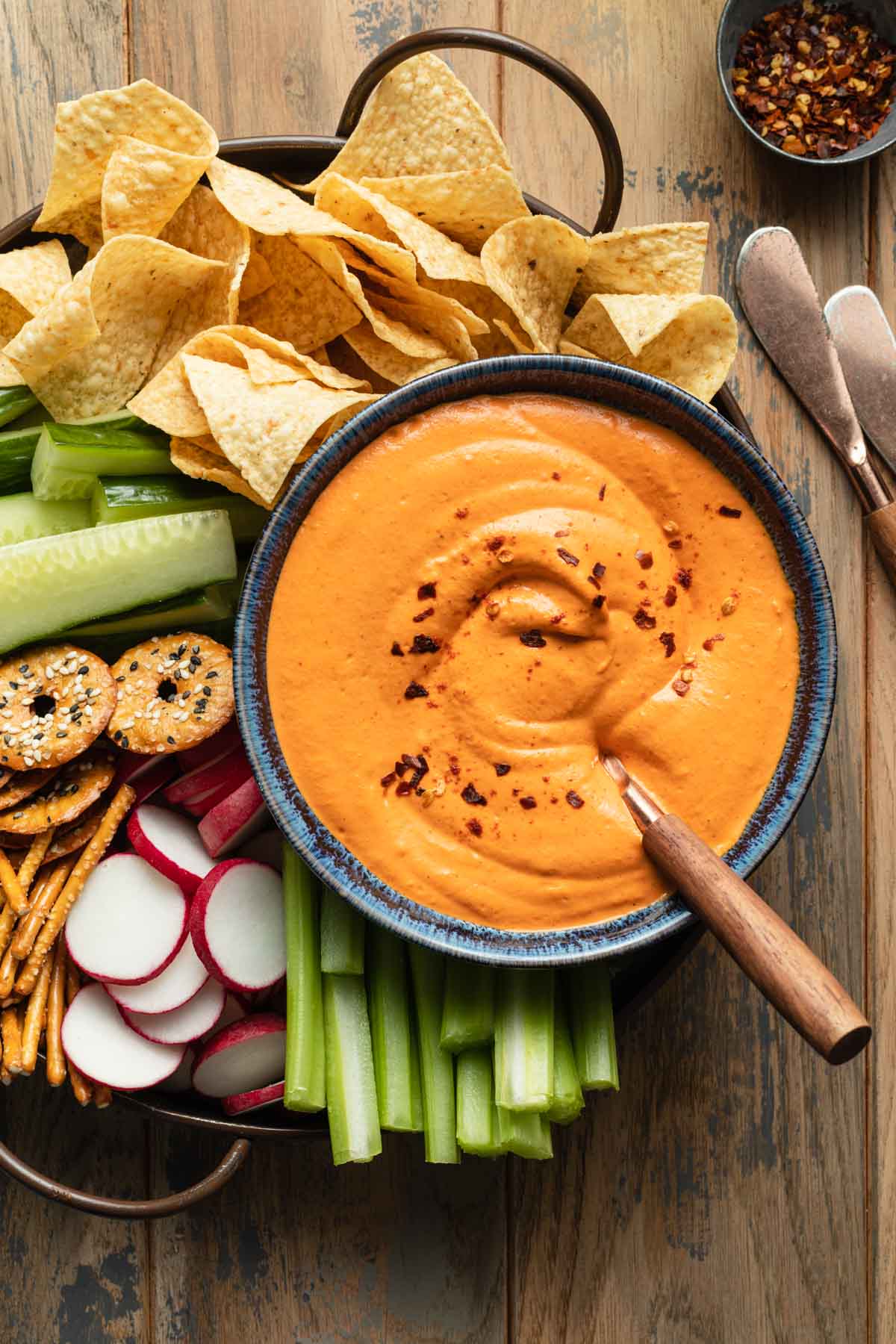 Vegan red pepper dip in a blue bowl on a tray with veggies and chips.