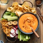 Overhead view of roasted red pepper dip served in a blue bowl with a tray of chopped veggies, pretzels and chips.