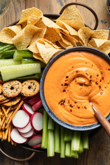 Overhead view of roasted red pepper dip served in a blue bowl with a tray of chopped veggies, pretzels and chips.