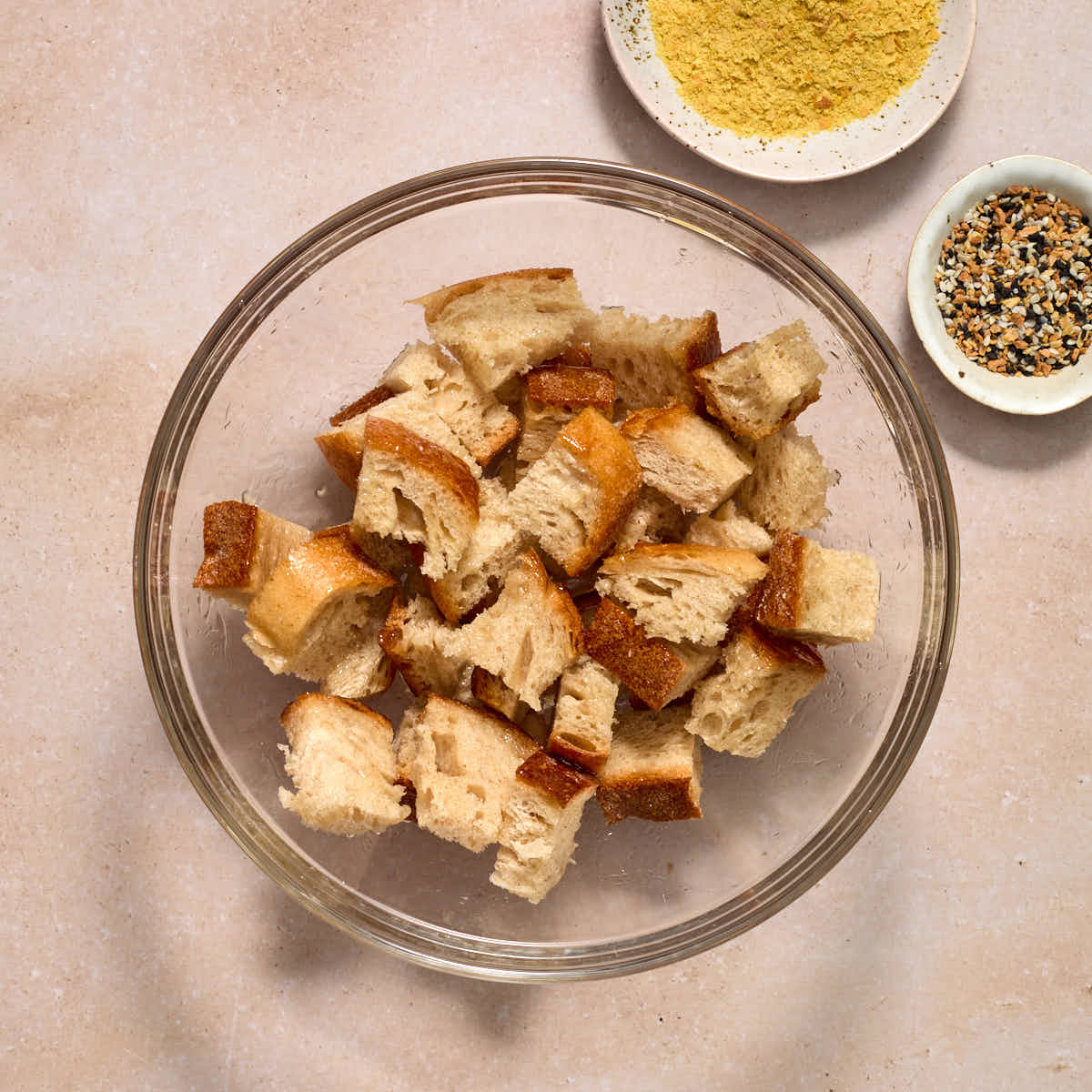 Bread cubes tossed in oil in a glass bowl with seasonings on the side.