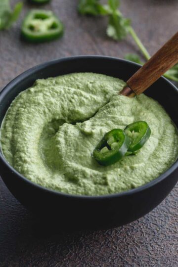 Angled view of creamy jalapeno dip in a small black bowl with jalapeno slices on top.