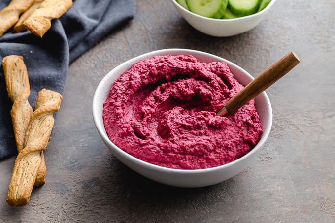Roasted beetroot hummus served in a white bowl with bread sticks on the side.