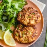 Two air fryer salmon patties on a plate with greens and lemon wedges.