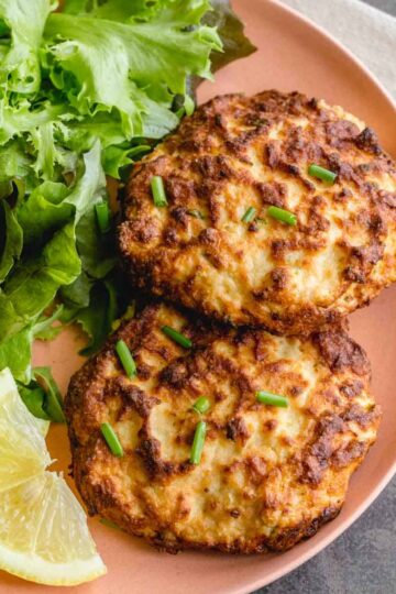Two air fryer salmon patties on a plate with greens and lemon wedges.