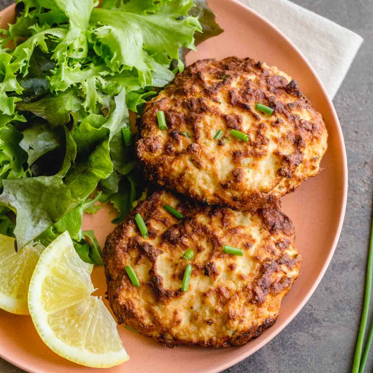 Two air fryer salmon patties on a plate with greens and lemon wedges.