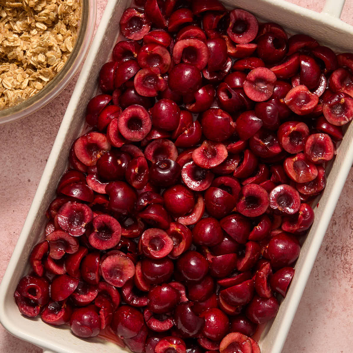 Cherries spread out in a white rectangular baking dish with bowl of crisp topping on the side.