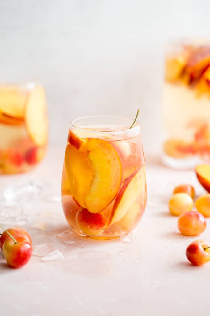 Side view of a glass of white wine spritzer with peaches and cherries.