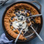 Overhead of a blueberry almond flour skillet cookie in a cast iron pan with spoons inserted and melted ice cream on top.
