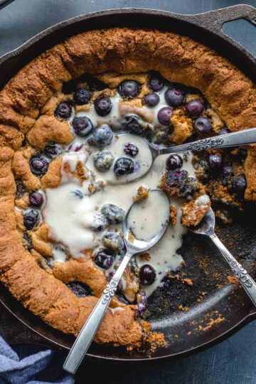 Overhead of a blueberry almond flour skillet cookie in a cast iron pan with spoons inserted and melted ice cream on top.
