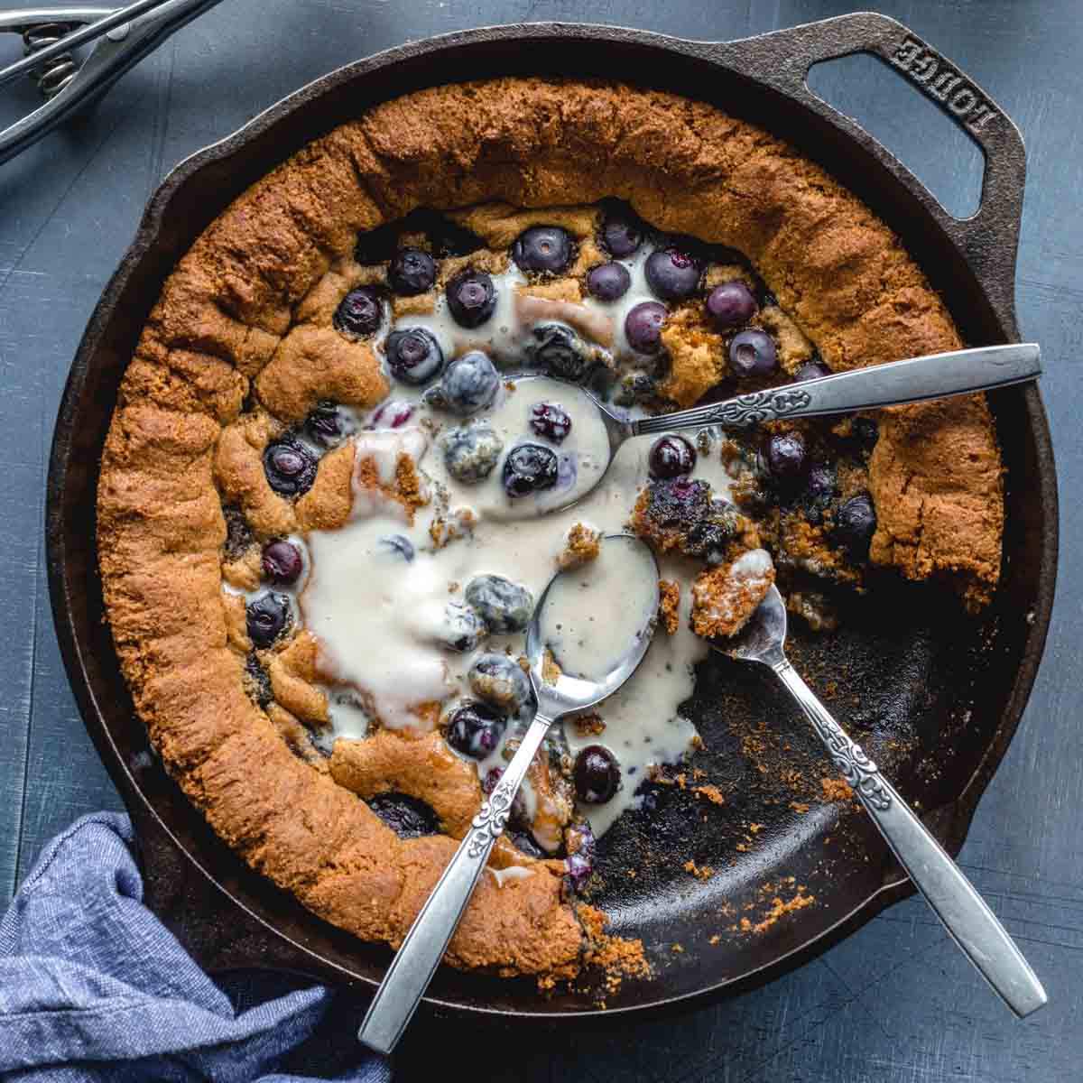 Overhead of a blueberry almond flour skillet cookie in a cast iron pan with spoons inserted and melted ice cream on top.
