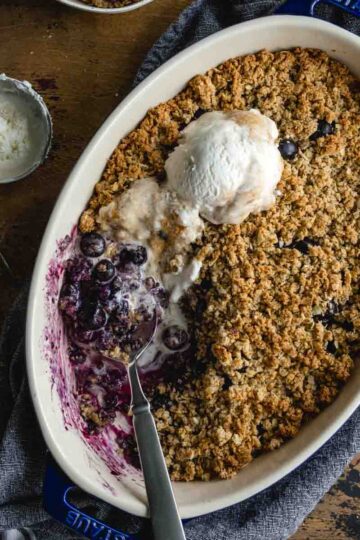 Overhead of a blueberry crisp in an oval dish with ice cream on top and some scooped out.